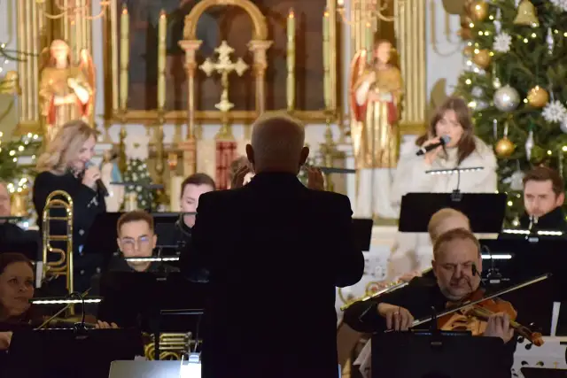 Dyrygent prowadzi orkiestrę i wokalistki w kościele, w tle choinka.