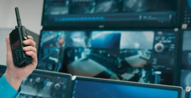 Dłoń trzyma krótkofalówkę na tle monitorów z podglądem z kamer, co sugeruje udostępnianie nagrań z monitoringu.