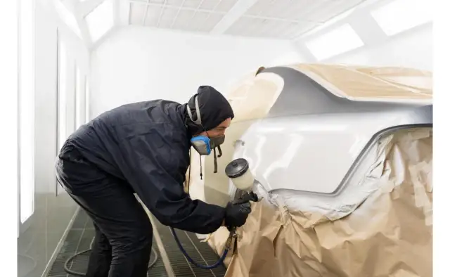 Lakiernik w masce i kombinezonie maluje samochód w kabinie. Czas schnięcia lakieru samochodowego zależy od wielu czynników.