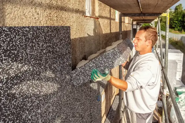 Pracownik montuje płyty izolacyjne na ścianie budynku. To etap ocieplenia domu, który zapewni komfort i oszczędność energii.