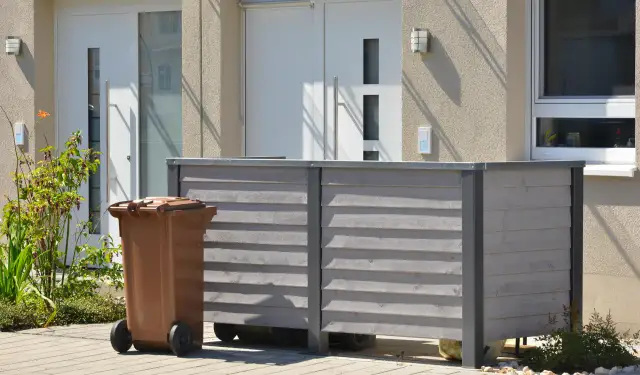 Nowy domek na śmieci to świetny sposób, jak ukryć kosz na śmieci w ogrodzie. Brązowy kosz stoi obok drewnianej obudowy.