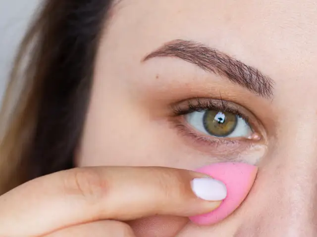 Zbliżenie na oko kobiety, która aplikuje korektor różową gąbeczką.