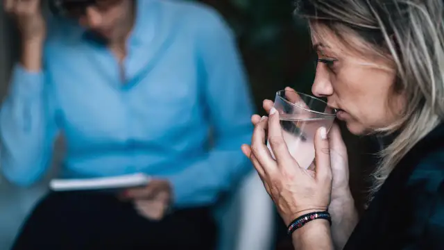 Kobieta pije wodę z kieliszka, siedząc naprzeciwko terapeuty.