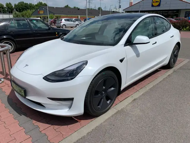 Biała Tesla Model 3 na parkingu przed sklepem. Zastanawiasz się, ile kosztuje najdroższa Tesla? Ten model to marzenie wielu kierowców.