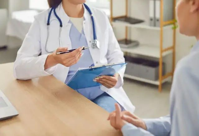 Lekarka w białym fartuchu z niebieskim stetoskopem rozmawia z pacjentem, trzymając teczkę.