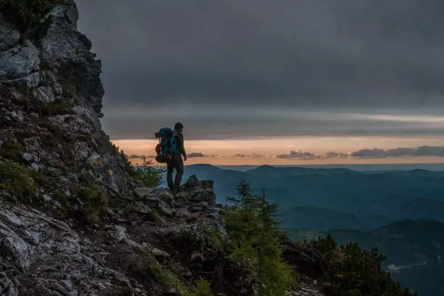 Turysta z plecakiem na skalnej półce, podziwiający zachód słońca nad pasmem górskim. Idealny widok, by zastanowić się, co zabrać w góry we wrześniu.