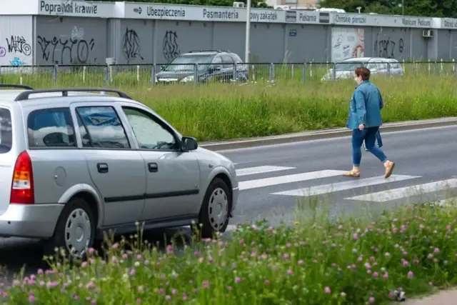 Kobieta w dżinsowej kurtce przechodzi przez przejście dla pieszych. Srebrny samochód czeka, a w tle widać budynki z napisami.