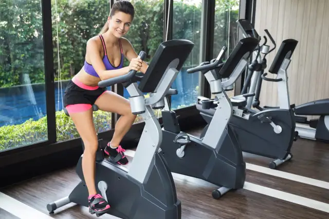 Uśmiechnięta kobieta ćwiczy na rowerze stacjonarnym. Dowiedz się, jak długo ćwiczyć na rowerze stacjonarnym, aby wspomóc kolana.