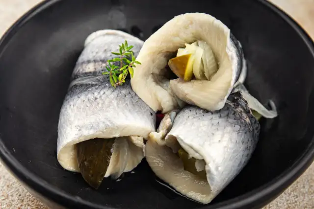 Roladki śledziowe z ogórkiem i cebulą, przygotowane przez moczenie śledzi w wodzie z octem, podane na czarnym talerzu.