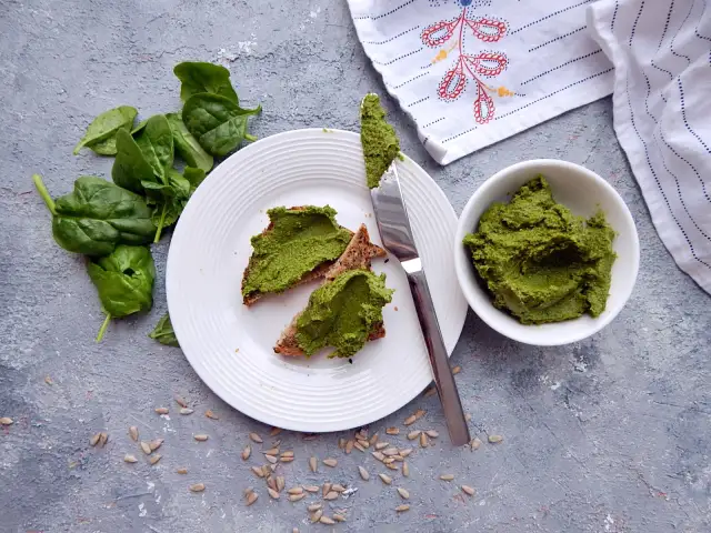 Pesto ze szpinaku do słoików na chlebie z pestkami słonecznika. Świeży szpinak obok talerza.