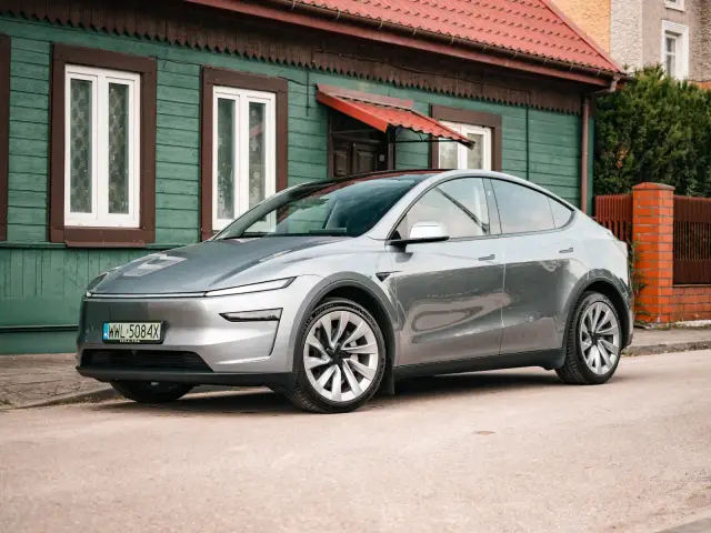 Srebrny Tesla Y stoi przed zielonym domem. Zastanawiasz się, ile koni ma ten model?
