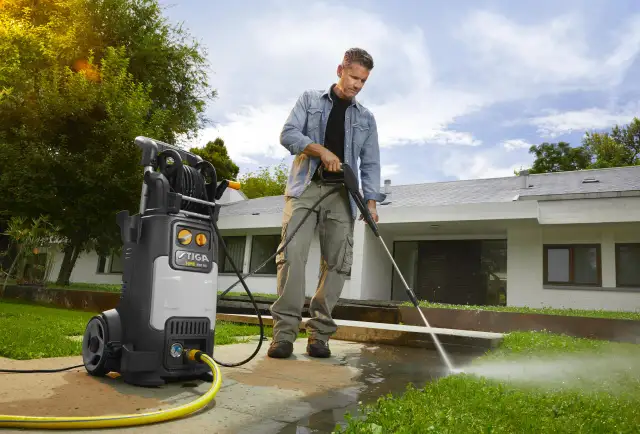 Mężczyzna myjką ciśnieniową czyści chodnik i trawę przed nowoczesnym domem.