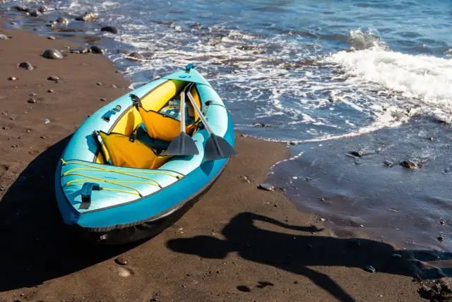 Niebiesko-żółty kajak pneumatyczny z wiosłami na piaszczystej plaży przy falującej wodzie.