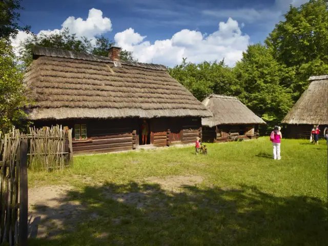 Drewniane chaty ze słomianymi dachami, trawiasty dziedziniec i ludzie.
