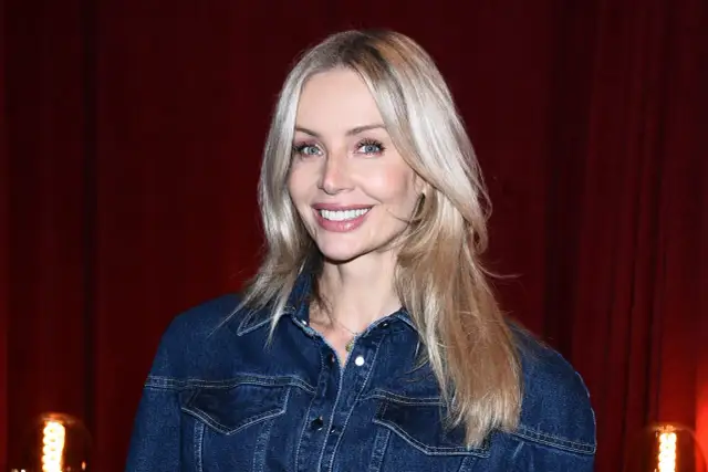 Uśmiechnięta blondynka z francuską grzywką w dżinsowej kurtce na tle czerwonej kurtyny.