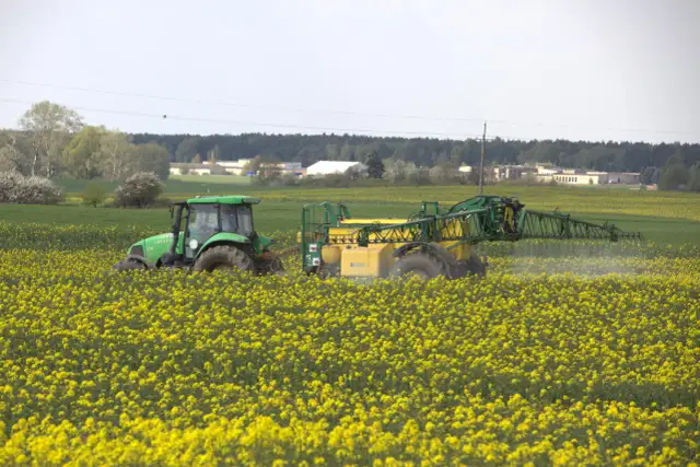 Zielony traktor z opryskiwaczem w polu rzepaku. Maszyna wykonuje zabieg mający na celu biologiczna ochrona upraw przed szkodnikami.
