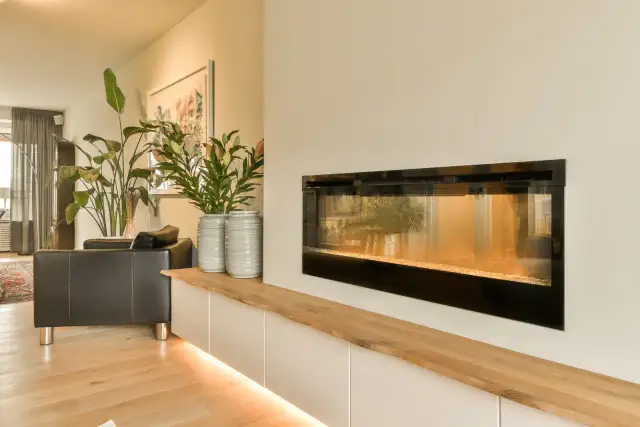 Nowoczesny salon z kominkiem, który można ozdobić, dodając rośliny i wazony. Drewniana półka nad szafkami podkreśla ciepło wnętrza.