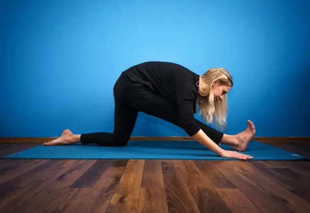 Kobieta w czarnym stroju ćwiczy rozciąganie na macie, wykonując pozycję przypominającą szpagat.