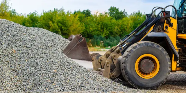 Ładowarka z zasypanym żwirem, widoczna z bliska, z zielonymi drzewami w tle.