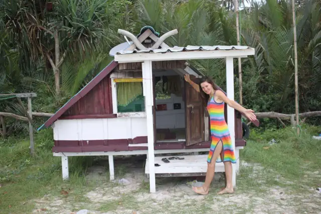 Kobieta w tęczowej sukience stoi obok małego drewnianego domku na plaży.