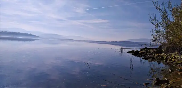 Spokojne jezioro z wędkami na brzegu, w tle góry i niebo z chmurami.