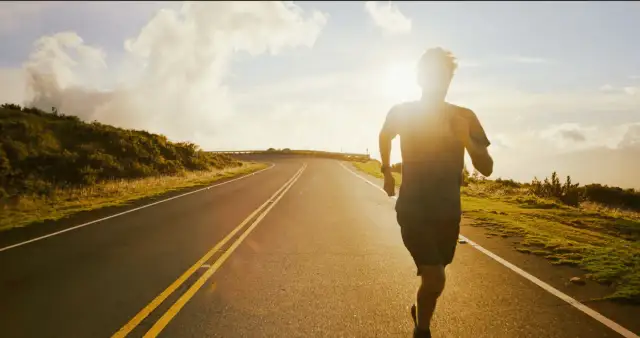 Sylwetka biegacza na tle zachodzącego słońca na pustej drodze.