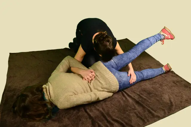 Dwie kobiety na brązowym kocu, jedna leży na plecach, druga klęczy nad nią, przykładając dłonie do jej bioder.