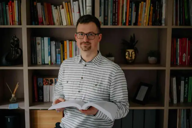 Mężczyzna w okularach, trzymający otwartą książkę, z neutralnym wyrazem twarzy, jakby uczył się, jak nie okazywać emocji na twarzy.