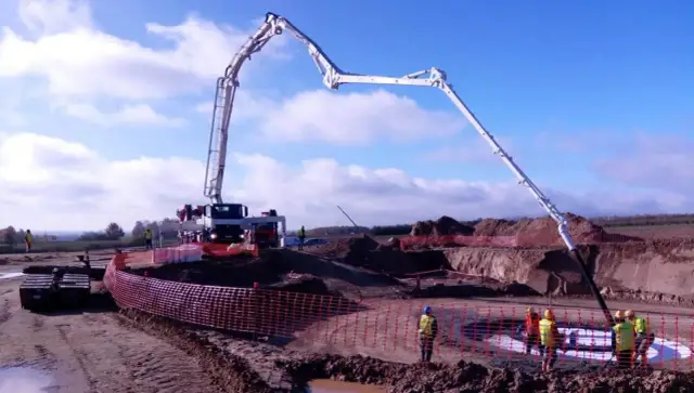 Ogromna pompa do betonu pracuje na budowie, dostarczając materiał do wykopu. Pracownicy w kamizelkach obserwują proces.