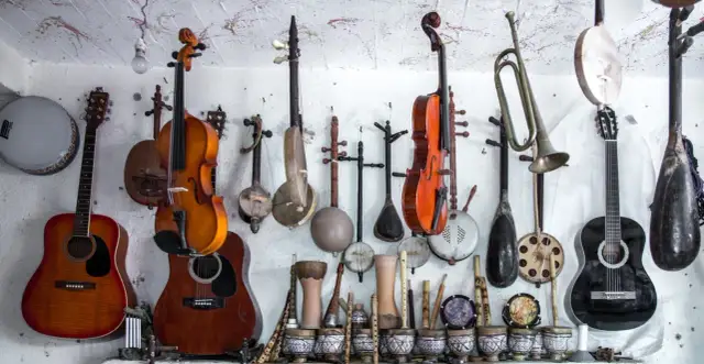 Różnorodne instrumenty strunowe, w tym gitary, skrzypce i tradycyjne lutnie, wiszą na ścianie obok bębnów i fletów.