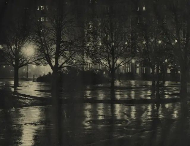 Nocny park po deszczu, drzewa bez liści, mokre ścieżki odbijające światła budynków. Alfred Stieglitz uchwycił atmosferę miasta.