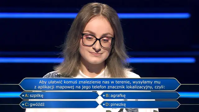 Młoda kobieta w okularach na tle niebieskich pasów, zastanawia się nad pytaniem z teleturnieju: "Aby ułatwić komuś znalezienie nas w terenie, wysyłamy mu z aplikacji mapowej na jego telefon znacznik lokalizacyjny, czyli: B: agrafkę".