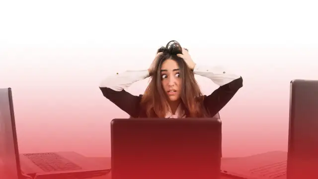 Kobieta z przerażeniem w oczach, rękami wplątanymi we włosy, siedzi przy biurku otoczona laptopami. To obraz stresu w pracy.