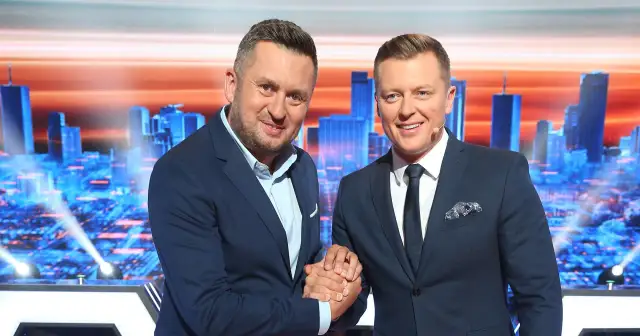 Dwóch mężczyzn w garniturach ściska sobie dłonie na tle scenografii z miejskim krajobrazem, gdzie nagrywane jest koło fortuny.