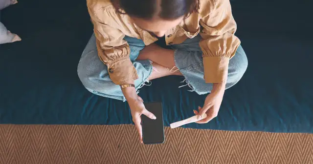 Kobieta siedzi ze skrzyżowanymi nogami, trzymając w dłoniach telefon i test ciążowy.