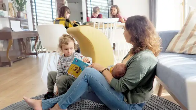 Mama karmi piersią niemowlę, podczas gdy starsze dziecko czyta książkę. To piękny przykład karmienia mieszanego w rodzinnej atmosferze.