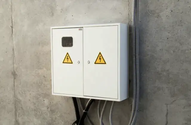 Biała skrzynka elektryczna z żółtymi symbolami ostrzegawczymi na betonowej ścianie.