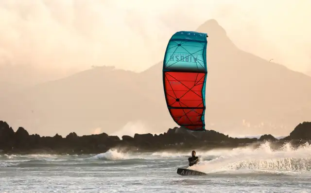 Kitesurfer na desce w akcji, z kolorowym latawcem na tle gór i morza.