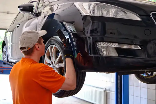 Mechanik w czapce i pomarańczowej koszulce ogląda podniesiony czarny samochód.