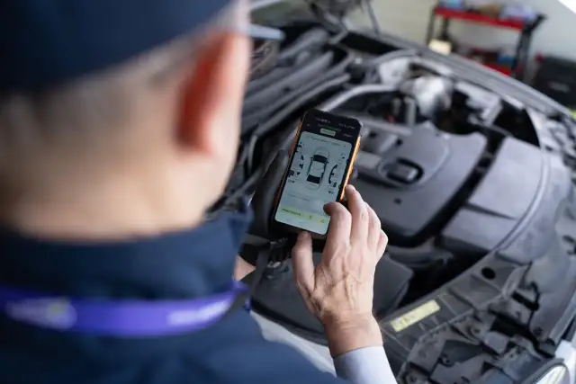 Mechanik w rękawiczkach trzyma smartfon z aplikacją do oceny stanu samochodu, na tle otwartej maski silnika.