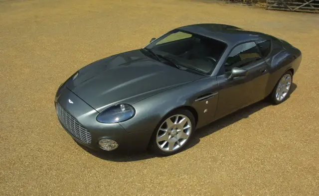 Szary Aston Martin DB9 na żwirowym podjeździe. Zastanawiasz się, ile kosztuje Aston Martin? To auto to marzenie wielu.