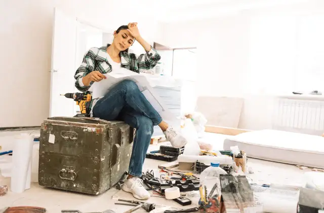 Zmęczona kobieta z planami w ręku siedzi na skrzyni w trakcie remontu.