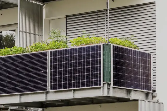 Panele fotowoltaiczne na balkonie, z zielonymi roślinami wyrastającymi ponad nimi. Ekologiczne rozwiązanie dla miejskiego ogrodu.