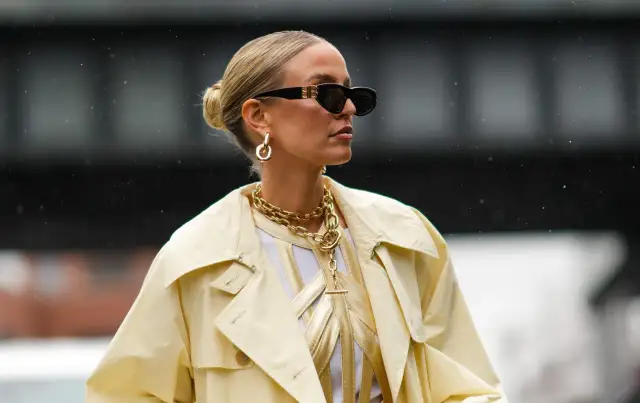Kobieta w okularach przeciwsłonecznych i żółtym płaszczu, z eleganckim upięciem włosów w kok. Złota biżuteria dodaje szyku.