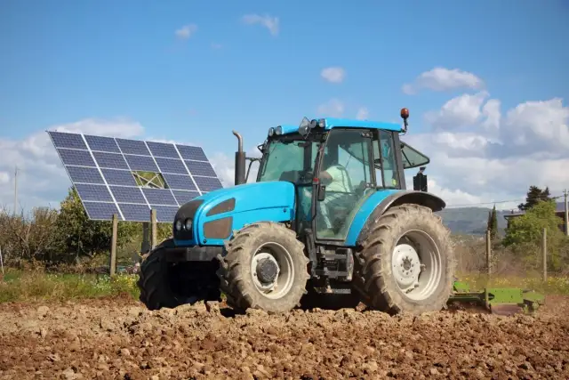 Błękitny traktor orze pole obok paneli fotowoltaicznych. Rolnik korzysta z dofinansowania dla rolników fotowoltaika, inwestując w zieloną energię.