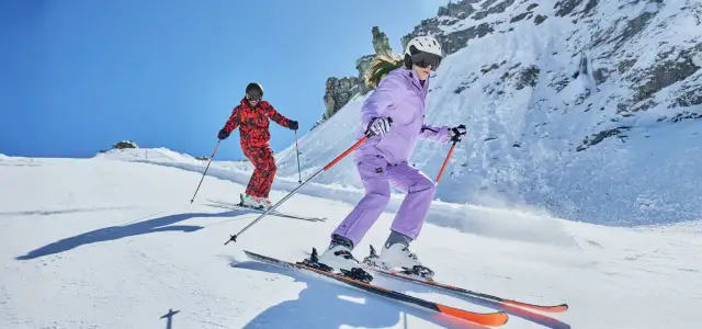 Dwoje narciarzy w kolorowych strojach zjeżdża po ośnieżonym stoku w słoneczny dzień.