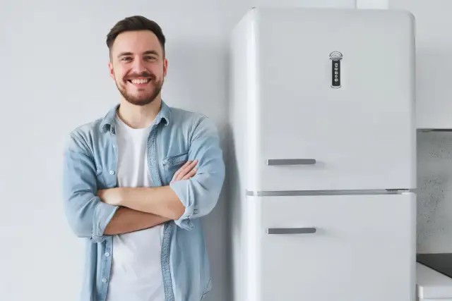 Uśmiechnięty mężczyzna w jeansowej koszuli stoi obok białej lodówki.