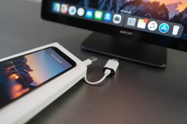 iPhone podłączony do komputera kablem, gotowy do zgrania zdjęć. Na ekranie komputera widać ikonki aplikacji.