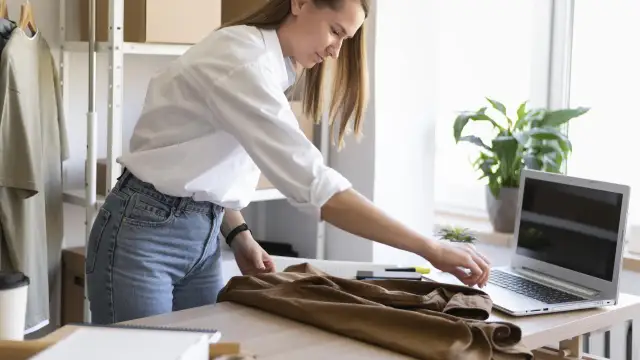 Kobieta w białej koszuli i jeansach mierzy brązową koszulę na biurku obok laptopa.