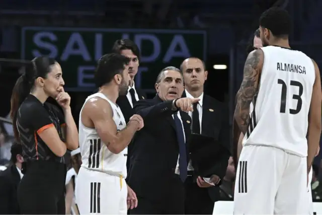 Entrenador del r madrid baloncesto señala una jugada. Jugadores y árbitra observan.